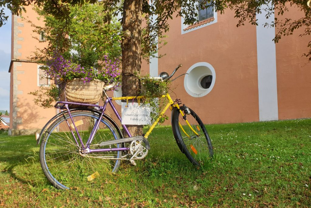 Mit Blumen geschmücktes Simultankirchen-Fahrrad in den Kirchenfarben gelb und violett vor St. Johannes Baptista in Floss
